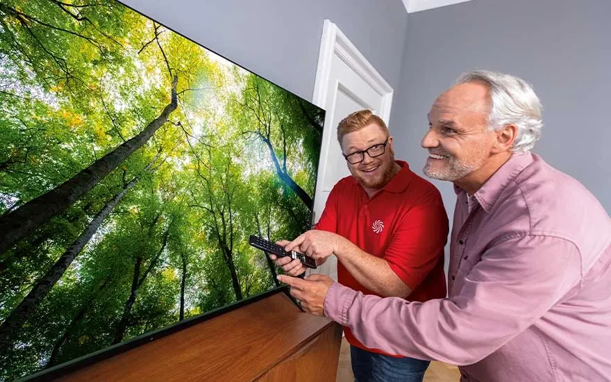 An in‑store expert helps an older customer set up a large modern TV. A bright forest with tall green trees fills the vivid screen.