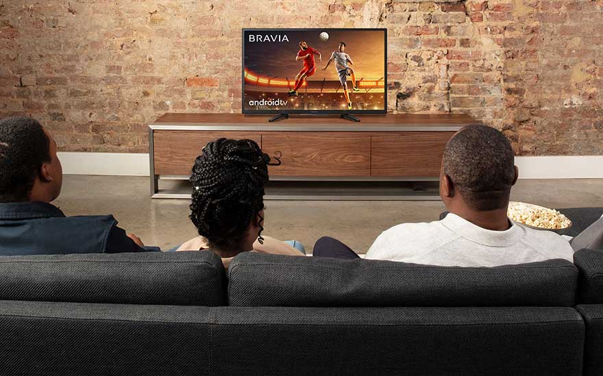 Three people sit on a sofa in a cosy room, watching a football match on a television placed against an exposed brick wall.