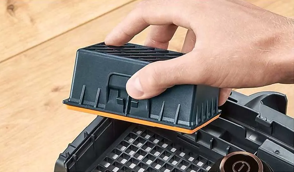 A hand inserts a black rectangular air filter with an orange edge into a device, onto a grid, on a wooden surface.