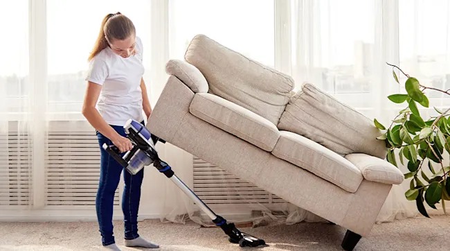 Vrouw stofzuigt onder een beige bank in een lichte kamer met een raam en een groene plant.