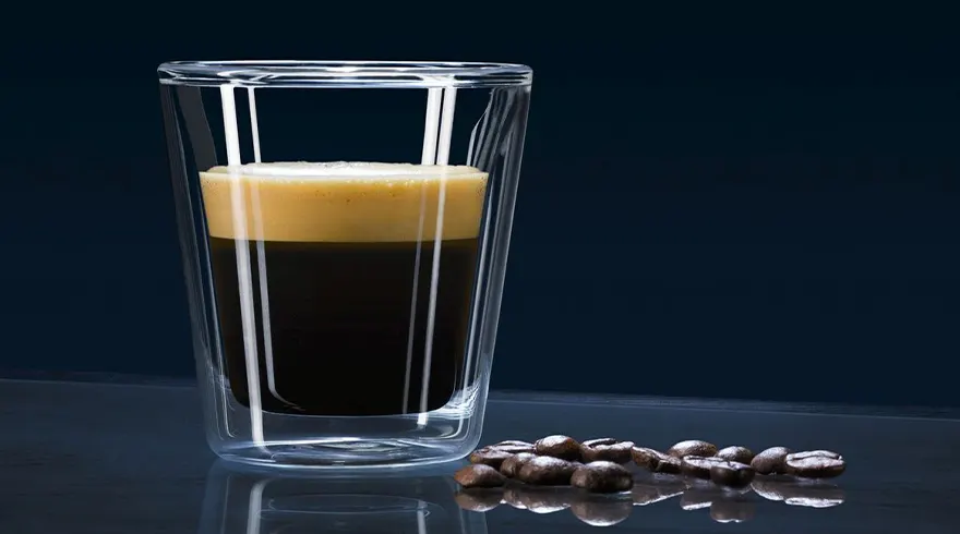 Een helder glas met koffie en een crèmelaag staat naast een stapel koffiebonen tegen een donkere achtergrond.