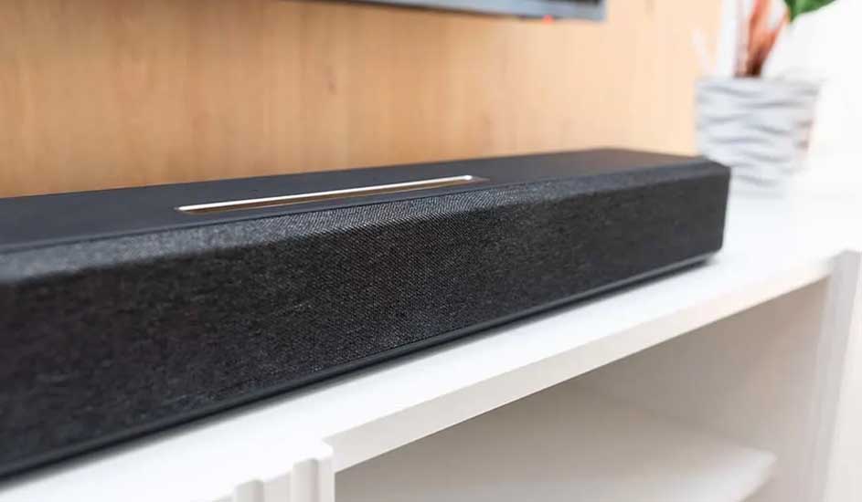 Dit is een zwarte soundbar met stoffen voorkant en een metalen detail bovenop, geplaatst op een witte kast in een moderne woonkamer.
