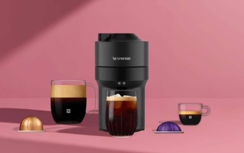 Zwarte Nespresso koffiemachine met twee glazen kopjes koffie en twee capsules op een roze achtergrond.