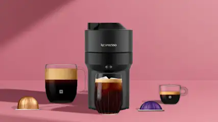 Zwarte Nespresso koffiemachine met twee glazen kopjes koffie en twee capsules op een roze achtergrond.