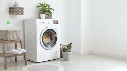 Wasmachine in een witte kamer, met planten en een kruk met slippers. Aan de muur een plank met een mand en een fles.