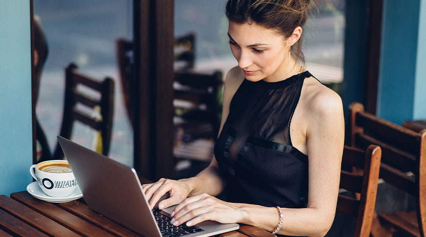 Vrouw typt op laptop aan houten tafel met koffie. Stoelen en blauwe muur op de achtergrond.