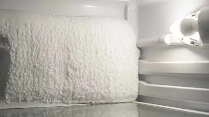 Een wit interieur van een vriezer met een dikke ijslaag op een compartiment, een lamp en plastic rekken.