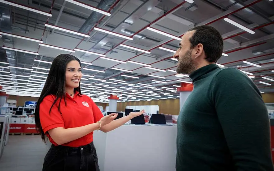 In een elektronicazaak verwelkomt een lachende medewerkster in rood poloshirt een klant tegenover haar, tussen uitgestalde laptops.