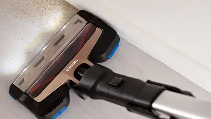 Een Philips stofzuiger met een zilveren kop en een zwarte buis zuigt vuil van een beige plint tegen een witte muur.