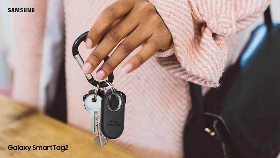 Een hand houdt een sleutelhanger met sleutels en een zwarte SmartTag tegen een achtergrond van een roze gestreepte trui en een zwarte tas.