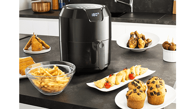 Op een aanrecht staat een zwarte airfryer omringd door borden met friet, snacks, muffins en gefrituurde kip.