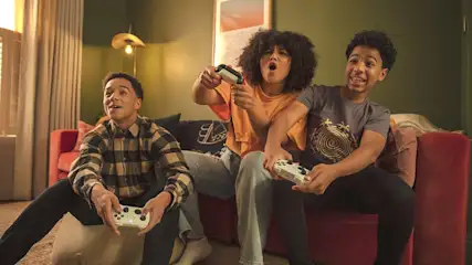 Drie jongeren zitten op een rode bank en spelen videogames met witte controllers.