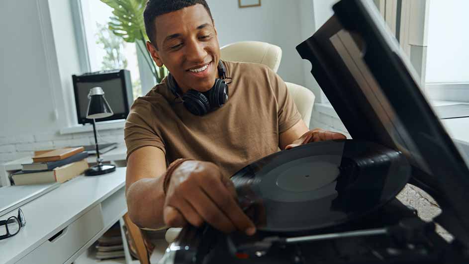 Een man met een koptelefoon plaatst een zwarte vinylplaat op een platenspeler. Op de achtergrond een bureau met boeken en een lamp.