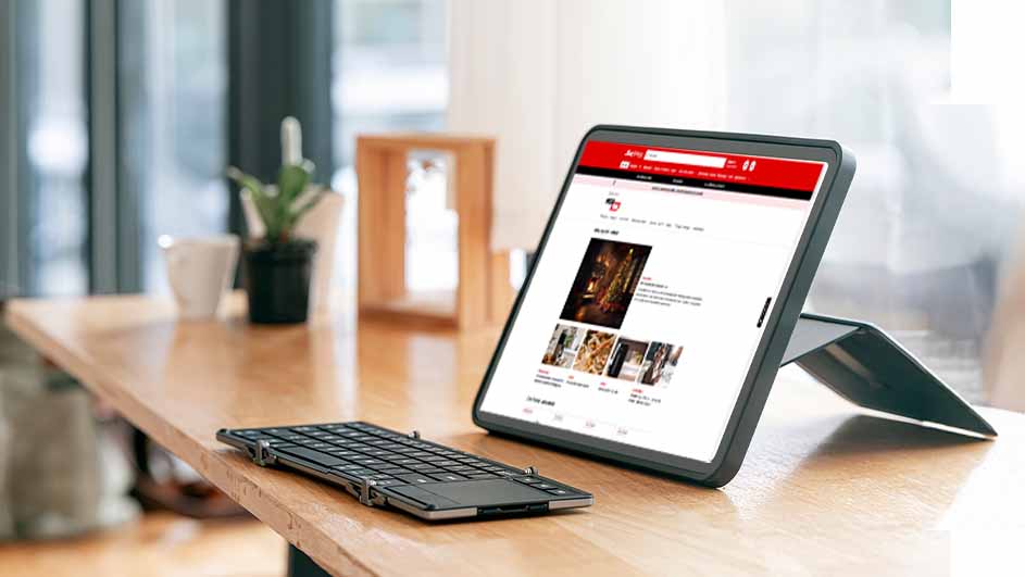 Een tablet met een rode website en een zwart toetsenbord op een houten tafel. Een plant en een mok zijn op de achtergrond zichtbaar.