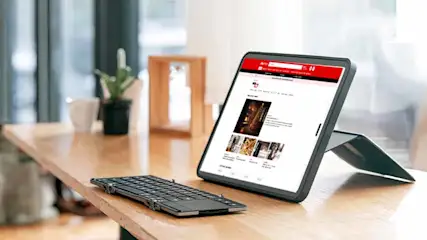 Een tablet met een rode website en een zwart toetsenbord op een houten tafel. Een plant en een mok zijn op de achtergrond zichtbaar.