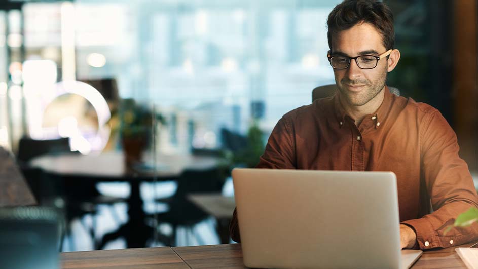 Een man met een bruin overhemd en een bril zit achter een zilverkleurige laptop aan een houten bureau in een kantoor.
