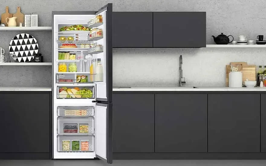 Built in fridge inside dark kitchen units stands open, revealing neatly arranged bottles, jars, vegetables and a cooking pot.