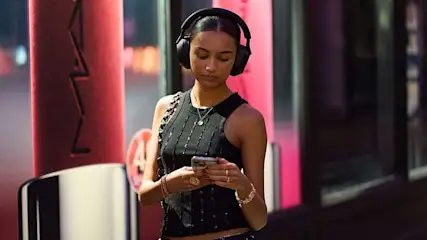 Een vrouw met zwarte draadloze Sony koptelefoon loopt buiten in een zwarte top, met een ketting en een tas over haar schouder.