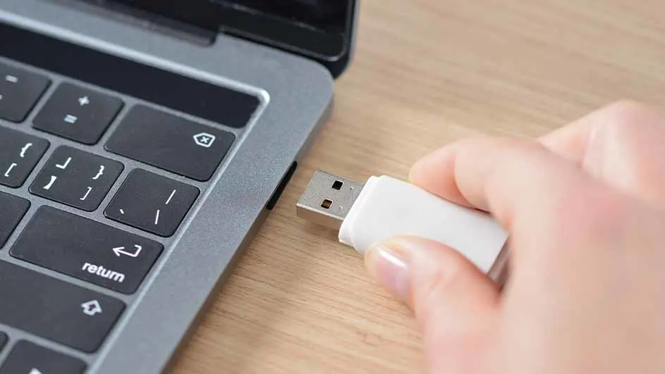 Een hand steekt een witte USB-stick in een grijze laptop op een houten tafel. Zwarte toetsen zijn zichtbaar op het toetsenbord.