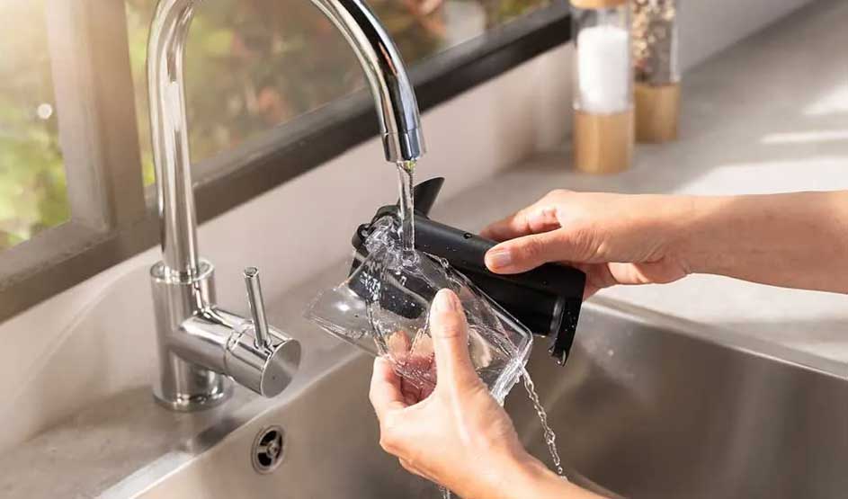 Je ziet handen die een melkopschuimer van het merk Philips onder een stromende keukenkraan afspoelen bij een moderne spoelbak.