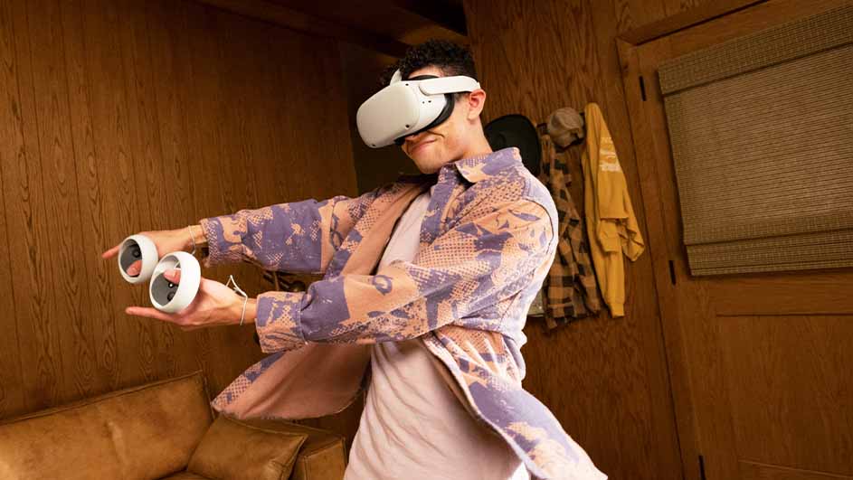 Man met VR-bril en controllers in een houten kamer. Kleding hangt aan de deur bij een bruine bank.