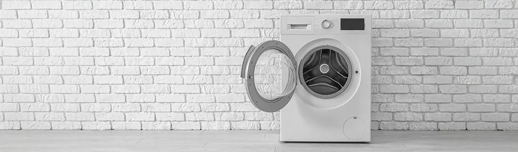 Een witte wasmachine met open deur staat tegen een witte bakstenen muur op een grijze vloer.