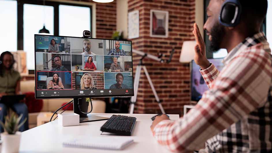 Een man met een koptelefoon zwaait naar een computerscherm met een videogesprek. Op de achtergrond een bakstenen muur.