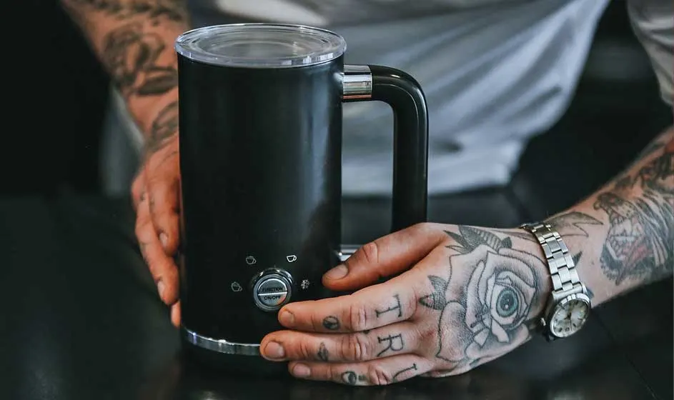 Getatoeëerde handen houden een zwarte melkopschuimer met handvat vast, op een donkere tafel, met knopjes onderaan zichtbaar.