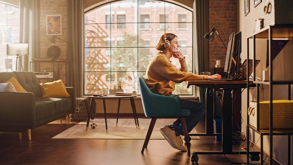 Vrouw met koptelefoon zit aan bureau met computer voor groot raam. Bank, tafel en kast in kamer.