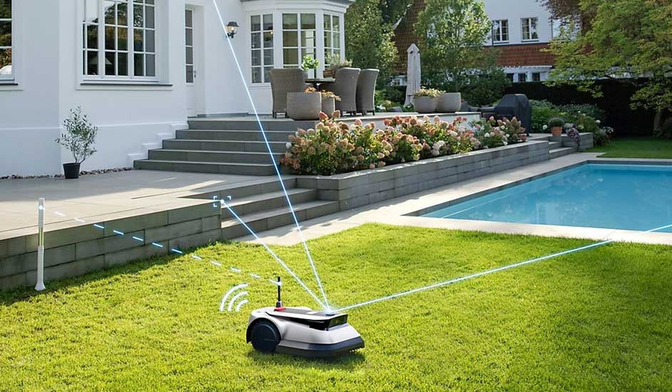 Een Gardena robotmaaier rijdt automatisch over het gras bij een modern huis met terras, bloemen en een zwembad op de achtergrond.
