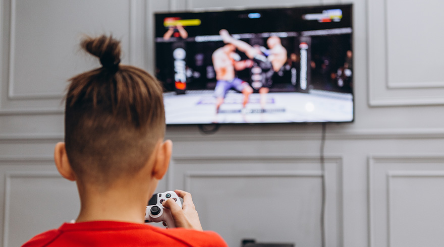 Een jongen in een rood shirt speelt een vechtspel op een tv aan een grijze wand. Hij gebruikt een witte controller.