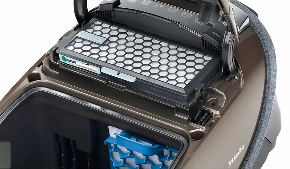 Een Miele stofzuiger met het deksel open toont een zeshoekig patroonfilter en een blauw voorfilter in het stofzuigercompartiment.
