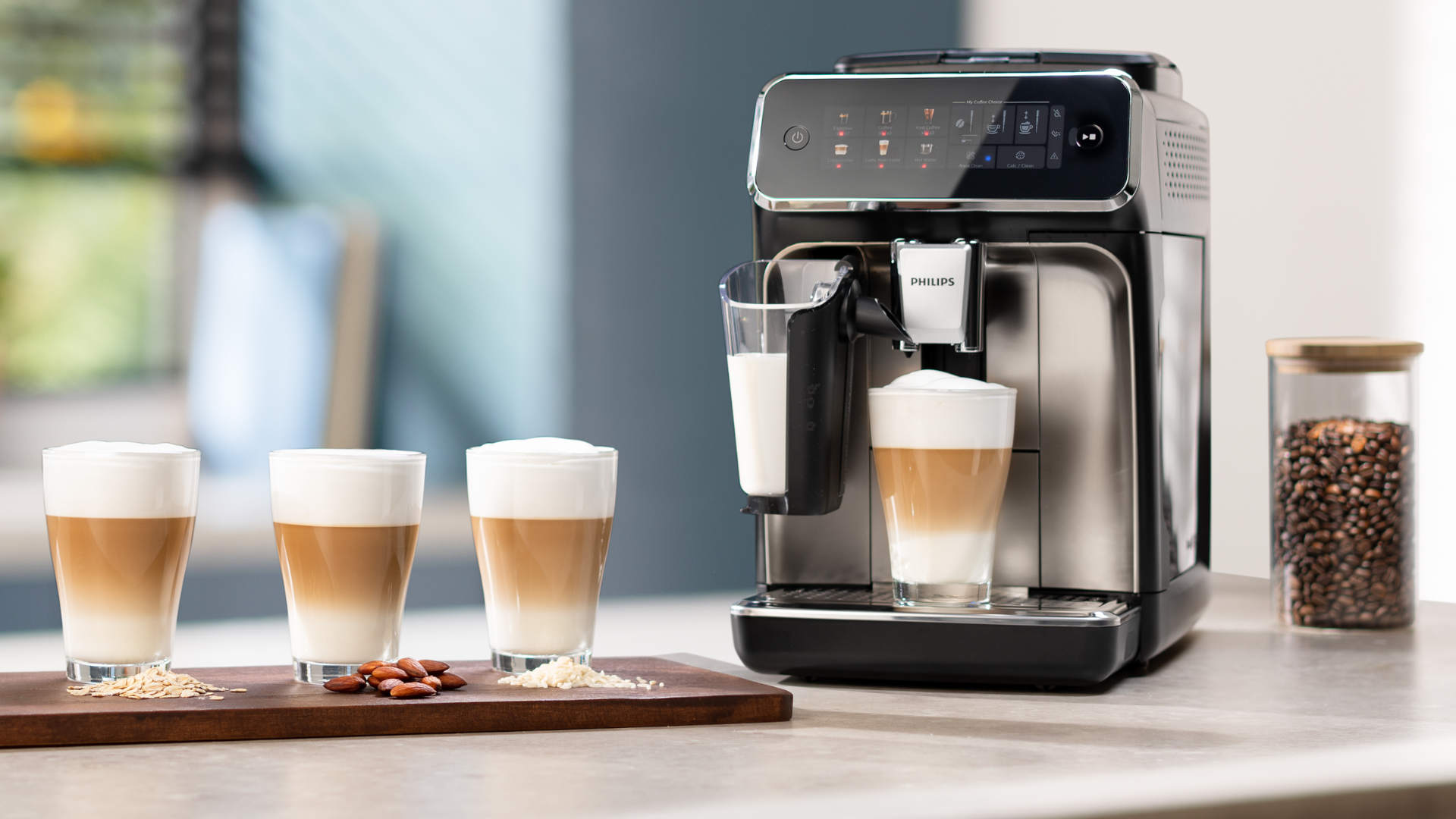 Koffiezetapparaat naast glazen latte macchiato op een houten plank, met een pot koffiebonen op de achtergrond.