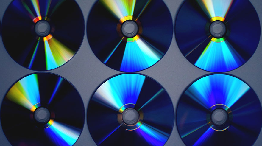 Zes cd's liggen op een rij tegen een grijze achtergrond, met reflecterende regenboogkleuren.