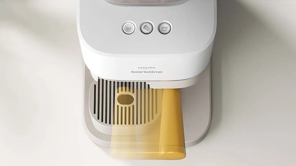 Een witte Philips Baristina-koffiemachine met drie knoppen en een gele beker eronder, tegen een lichte achtergrond.