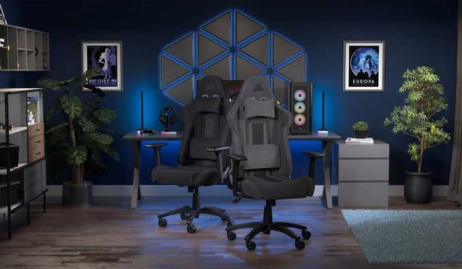 Two grey racing gaming chairs face a dual-PC desk in a dark blue room with neon wall panels, plants and sci‑fi posters.