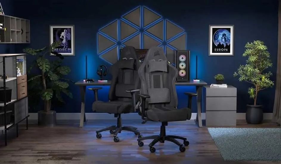 Two grey racing gaming chairs face a dual-PC desk in a dark blue room with neon wall panels, plants and sci‑fi posters.