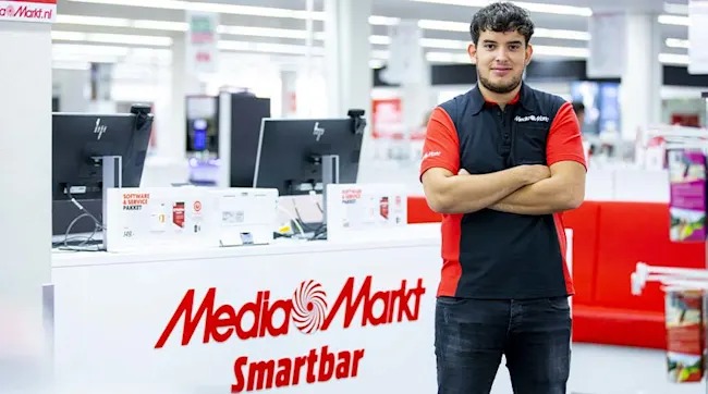 Een man in een rood en zwart Media Markt-shirt staat voor een Smartbar met computers en productdozen.
