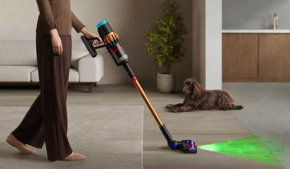 Een vrouw stofzuigt met een Dyson steelstofzuiger, groene laser toont stof op de vloer terwijl haar bruine hond ontspannen toekijkt.