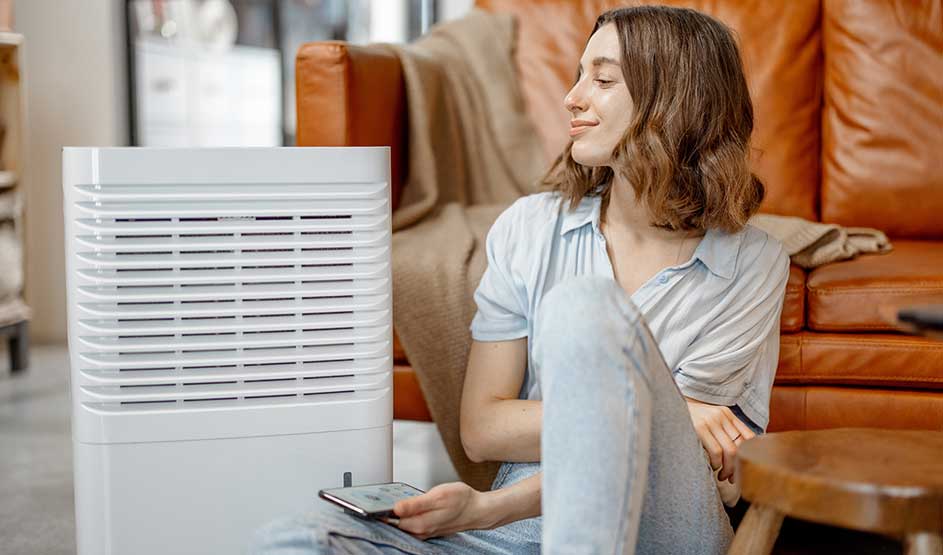 Een vrouw zit ontspannen op de vloer tegen een bruine leren sofa, glimlachend naar een witte luchtreiniger naast haar terwijl ze een smartphone vasthoudt.