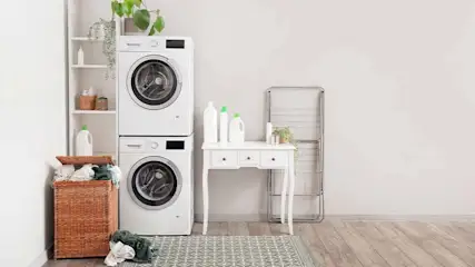 Laundry room with stacked white washer/dryer, wicker basket, white shelf, table, and folding rack on a patterned rug.