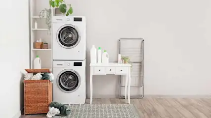 Een wasruimte met twee witte wasmachines op elkaar, een witte tafel, een rek en een bruine wasmand.