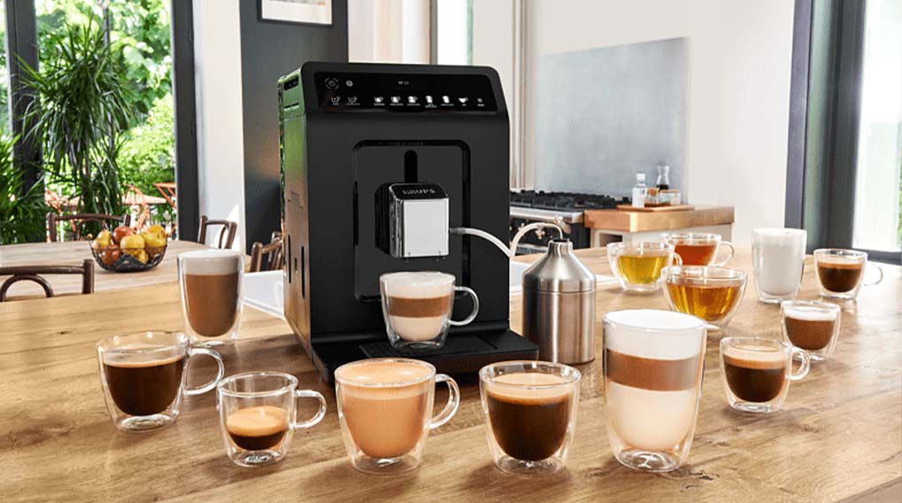 Zwarte koffiemachine met veel glazen bekers op een houten tafel. Achtergrond met raam, planten, en keuken.