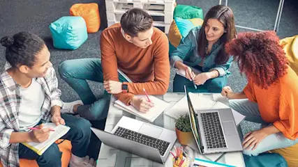 Vier personen zitten rond een tafel met laptops en notitieblokken. Kleurrijke zitzakken en een witte pallet op de achtergrond.