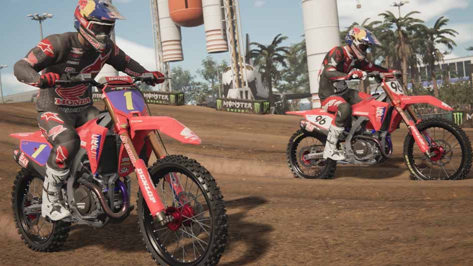 Twee motorcrossers op rode motoren rijden op een zandpad met palmbomen en reclameborden op de achtergrond.