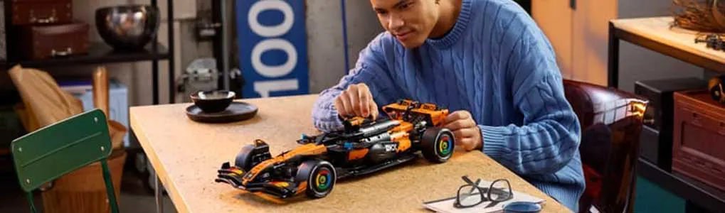 Man in blauwe trui bouwt aan een oranje LEGO Formule 1-auto op tafel, met bril, koffiekop en gezellige kamer eromheen.