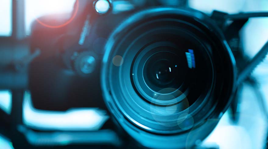 Close-up van een camera met een grote lens, in blauwtinten, met een onscherpe achtergrond en lichteffecten.