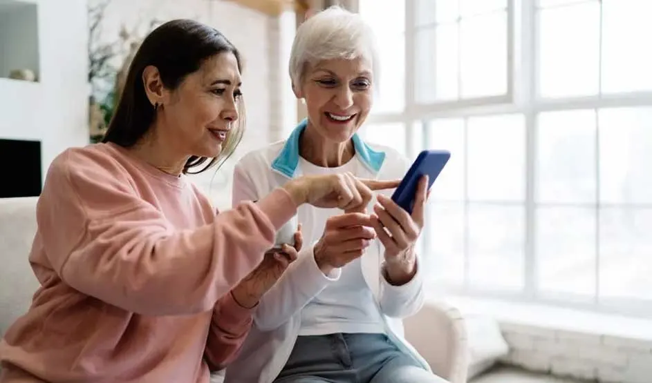 Two women sit together on a sofa, smiling as they look at a smartphone. One points at the screen, helping the other. No brand is visible.
