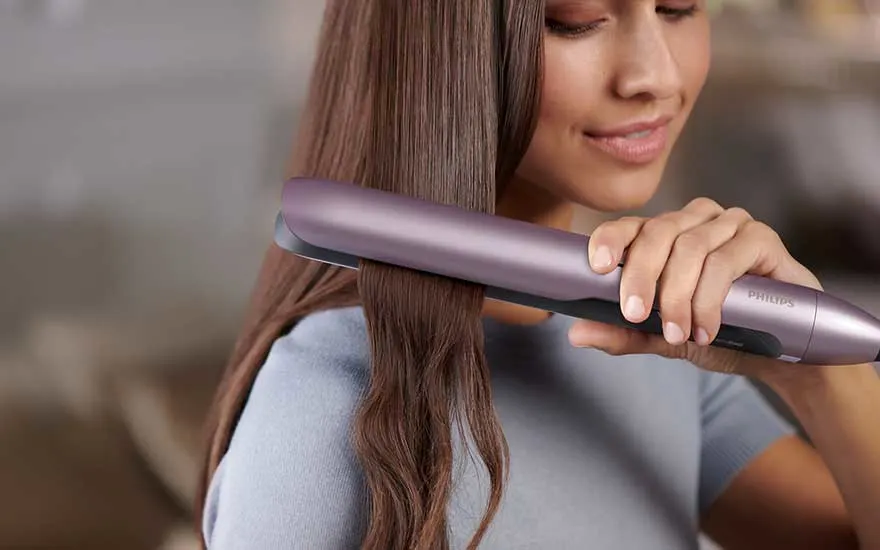 A smiling woman uses a purple Philips straightener to make her long brown hair look smooth, tidy and glossy at home.