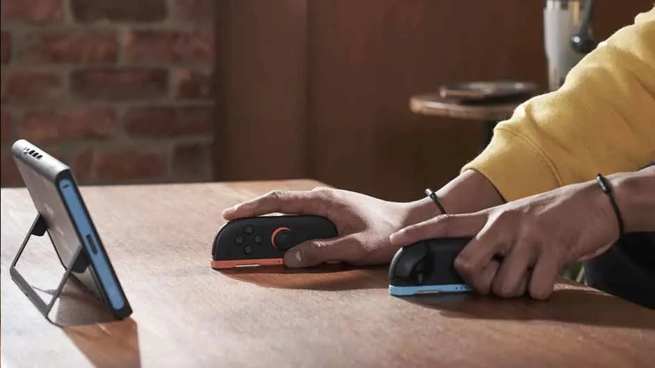 Een zwarte tablet staat op een houten tafel, naast twee handen die controllers vasthouden.
